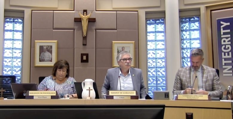 Three people sit at a table behind microphones. There is a small cross perched on the table and a large cross and picture of the pope on the wall behind them.