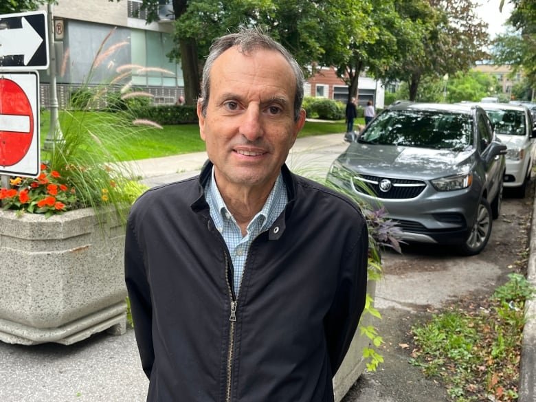 A man with very short grey hair smiles at the camera. He is wearing a light-coloured collared shirt under a dark zip-up jacket and holds his hands behind his back. Behind him is a residential street lined by trees and parked cars.