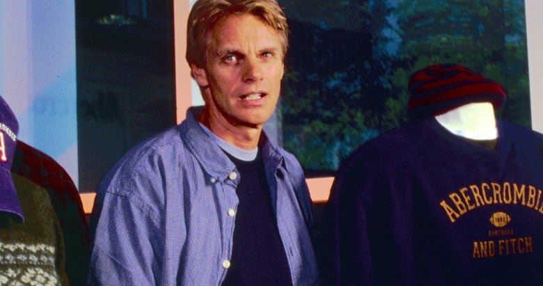 An older photo shows a man in a blue un-buttoned button-up, standing in between two mannequins, one of which is wearing a sweater that says Abercrombie & Fitch on it.