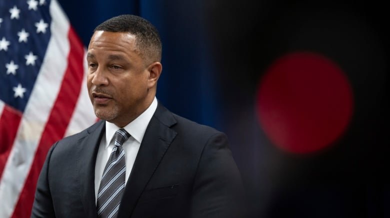 A man is shown from the chest up, wearing a suit and in the middle of talking. He is standing in front of a blue curtain wall and there's an American flag visible behind him as well.