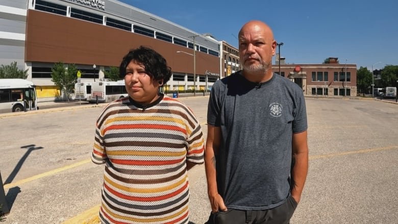 A young woman with short dark curly hair stands in an empty parking lot, facing straight on to the camera. The sky is clear and it is sunny. She is holding her hands behind her back and wears a striped short sleeved sweater. Her facial expression is neutral. To her left, stands a bald older man with a grey beard wearing a grey t-shirt. He is frowning slightly and has his hands in his pants pockets.