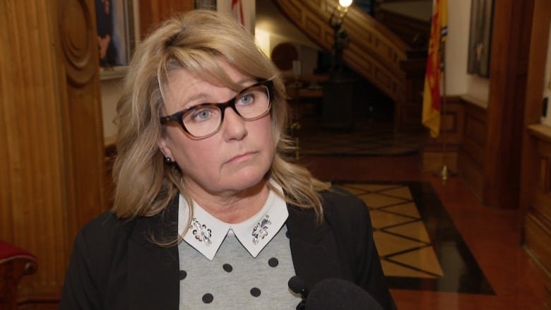 An unsmiling woman wearing a polka-dotted shirt with a bejeweled collar. She has blond shoulder-length hair and is wearing black glasses.