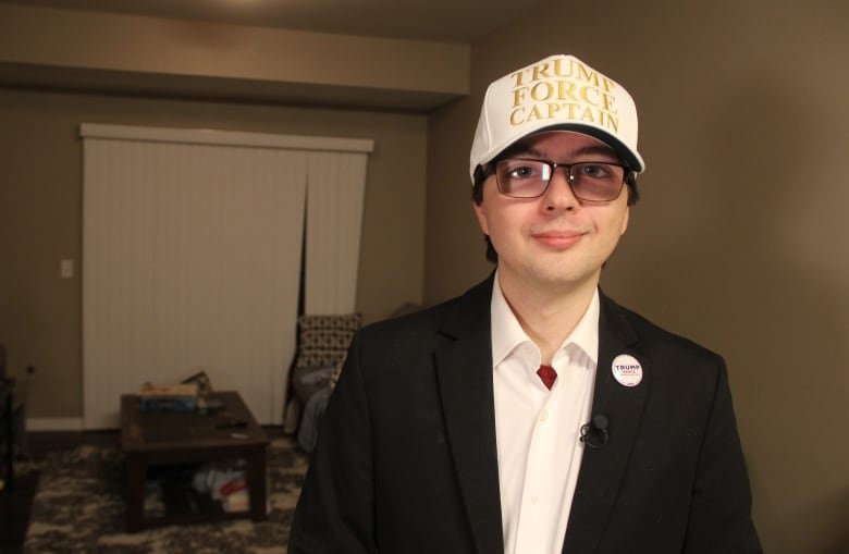 A man in a black blazer and white buttoned-up shirt underneath stands in a living room, while wearing a white hat with the letters in gold reading, "Trump Force Captain."