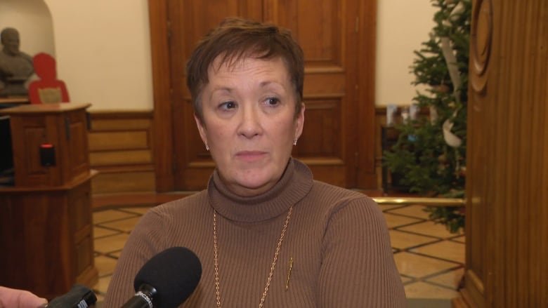 An unsmiling woman in a brown turtleneck standing in front of a black microphone