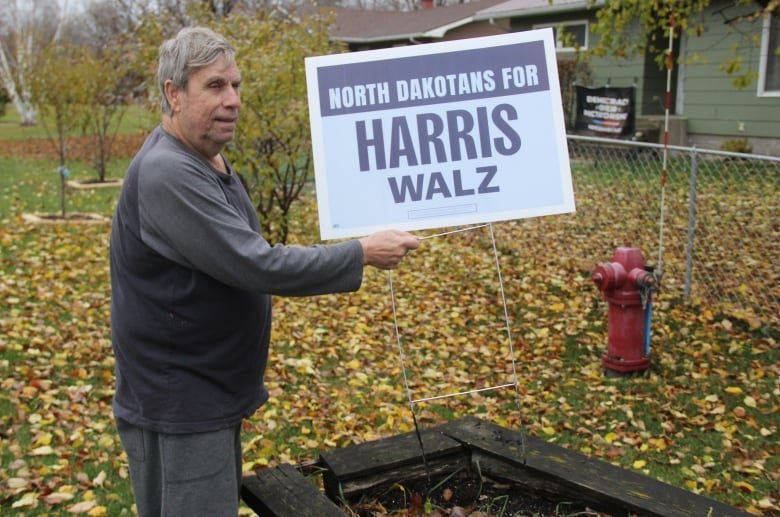 A man in a black and grey sweater holds a light blue sign that reads "North Dakotans for Harris Walz."