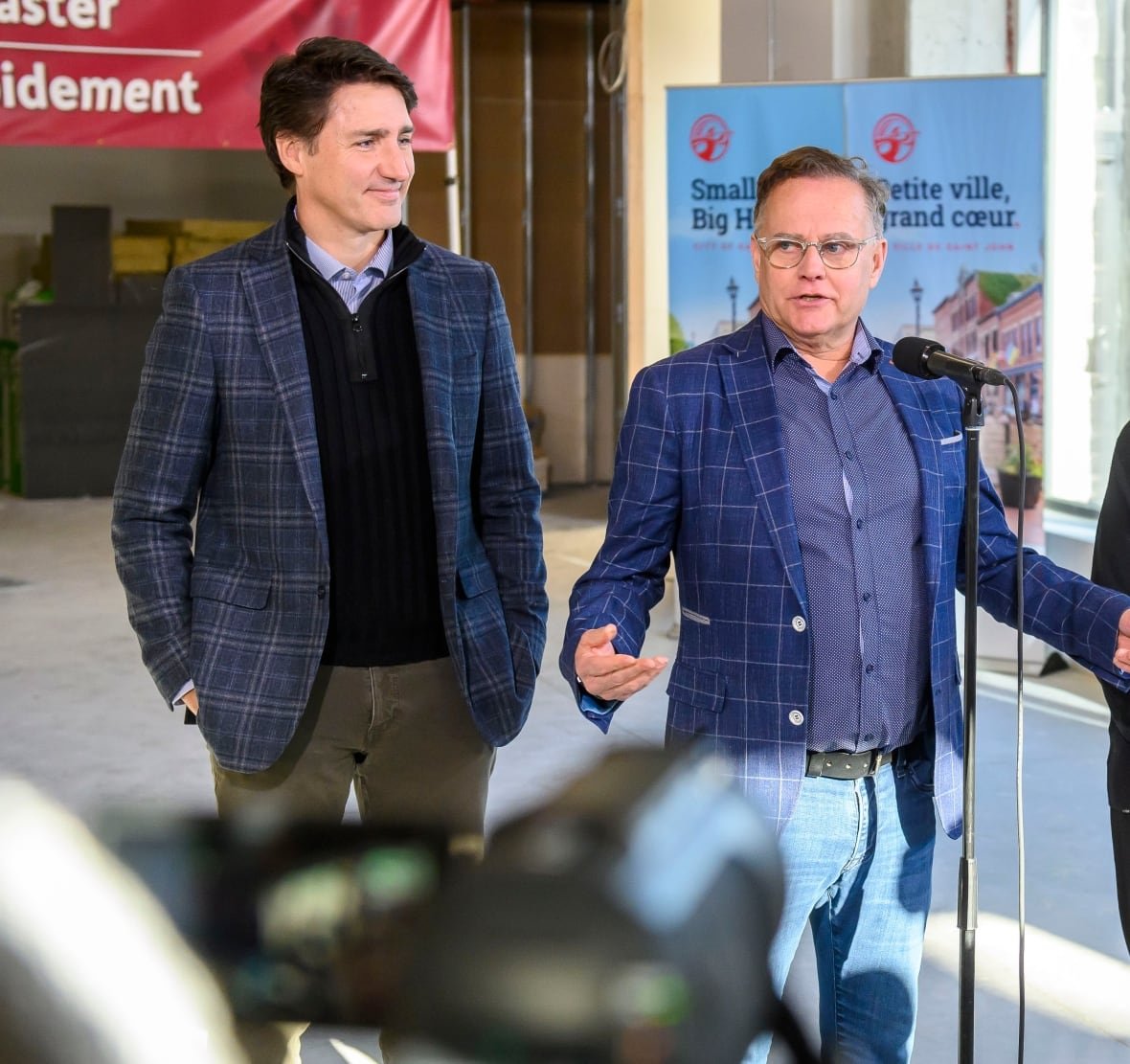 A brown-haired man and a grey-haired man, both wearing blue plaid sport jackets, stand in front of a microphone.