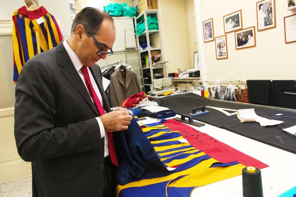 A man in a grey suit and red tie stitches material while standing in front of a work bench covered in coloured material.