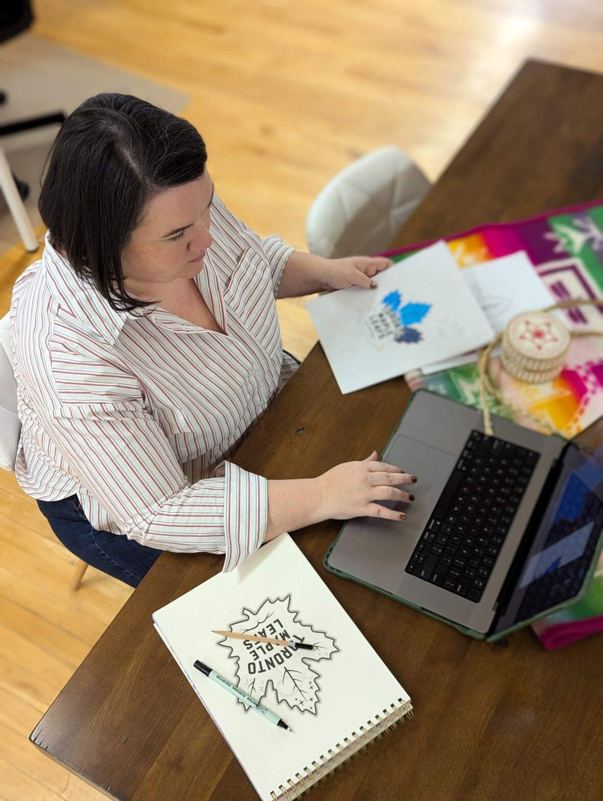 jennifer-taback-design A woman working on a laptop with a hand drawn image of a maple leaf on a table.
