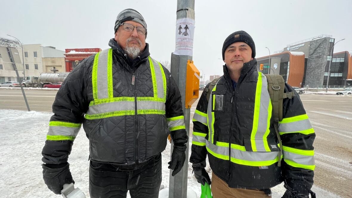 darryl-contois-and-george-robinson Two men wearing reflective vests stand on a snowy sidewalk on Main Street. A poster is taped to a light pole behind them.