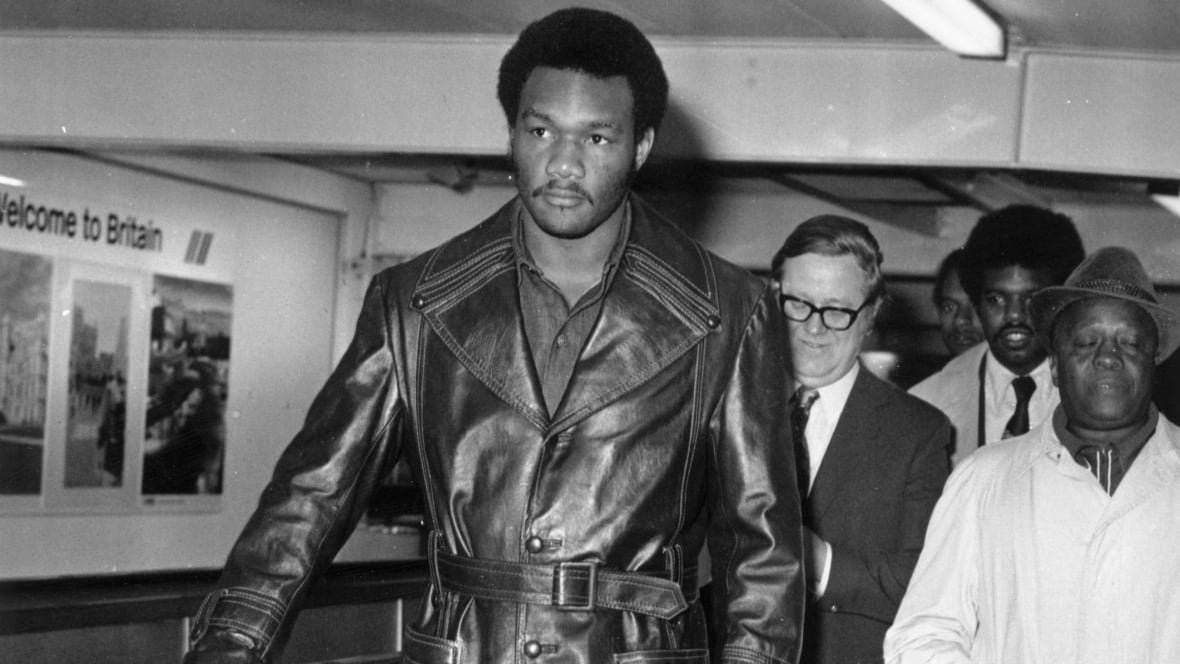 foreman-1973 A dark complected man is shown walking in a leather coat in a black and white photograph that appears to be decades old.