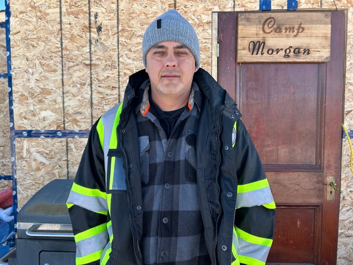 george-robinson Man wearing a grey tuque stands wearing an open jacket with reflective stripes. A sign behind him says Camp Morgan