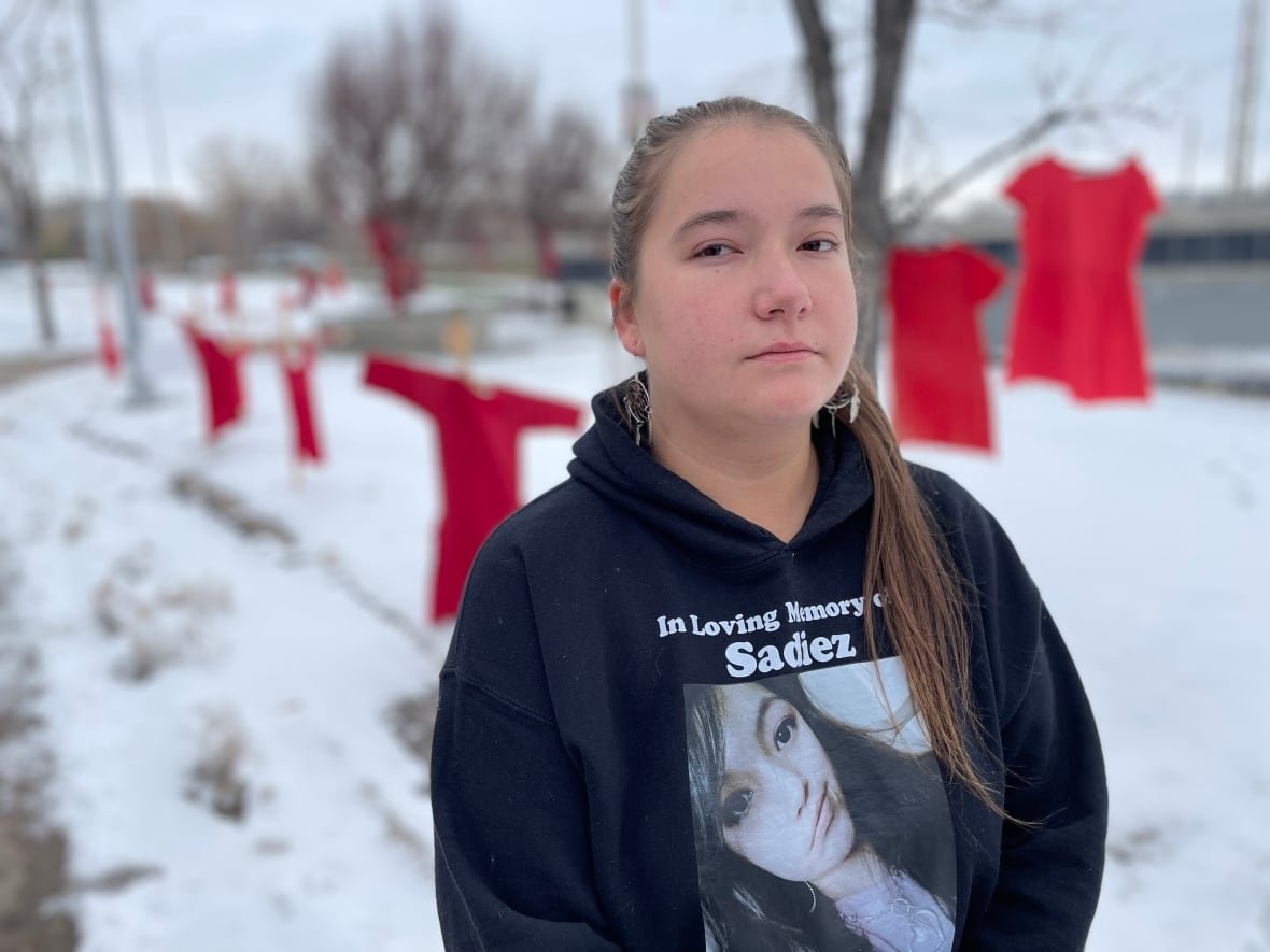 jorden-myran A girl in a black hoodie with a woman's photo and the words "in loving memory of Sadiez" on it stands outside.