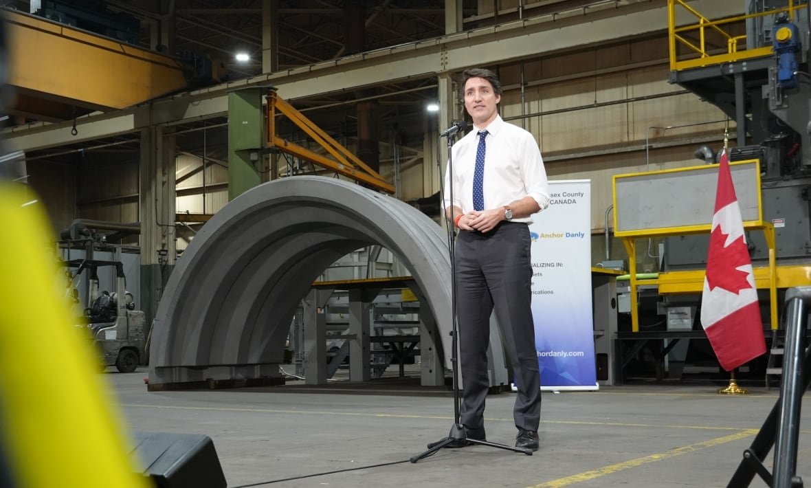 justin-trudeau-2 Trudeau standing at a microphone in a factor with what appears to be a large piece of concrete pipe behind him.