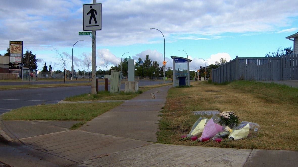 memorial-to-assault-victim Bundles of flowers lying on grass beside a sidewalk, with a pedestrian crosswalk and bus shelter in the background.