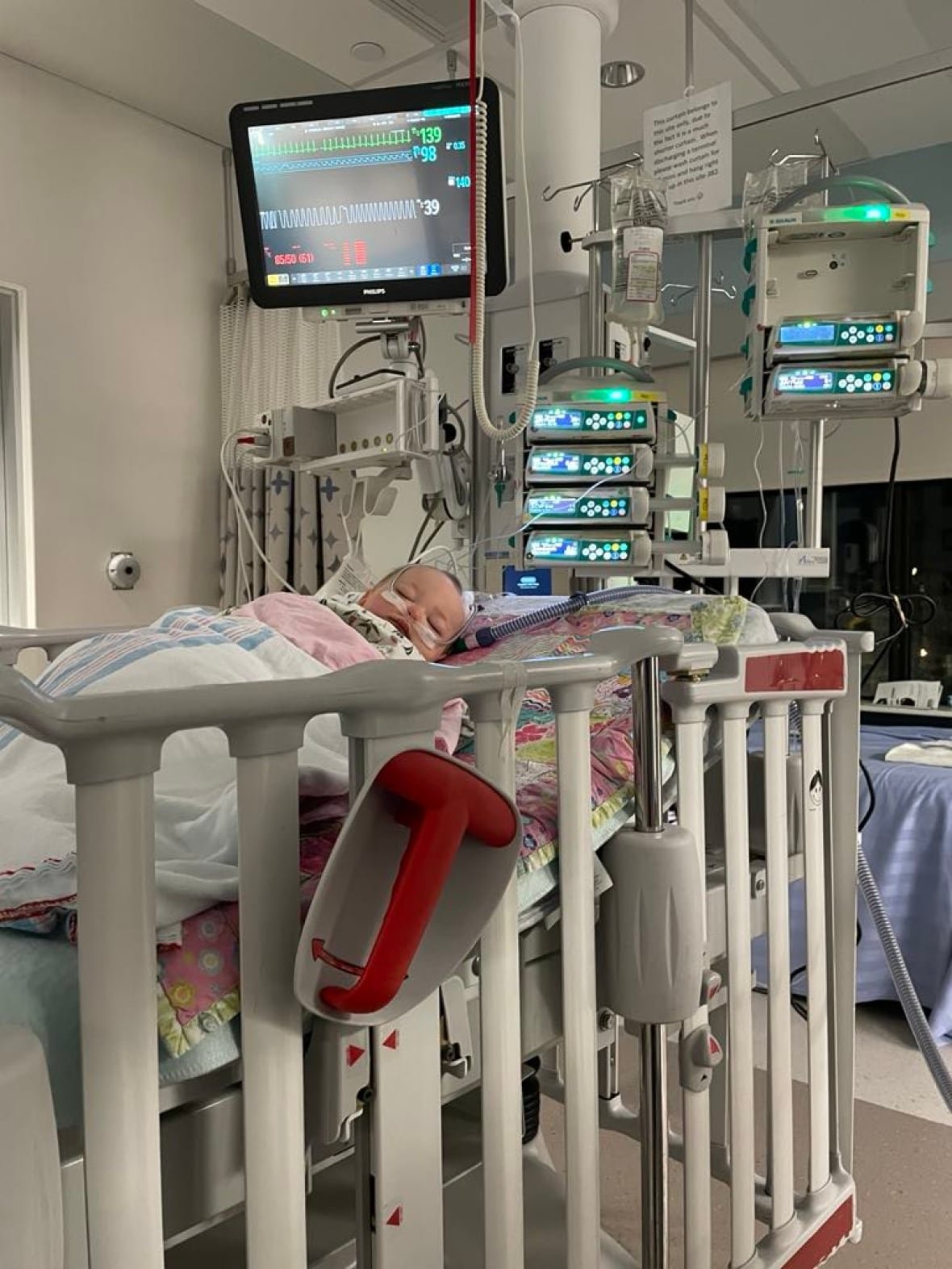 rosemary-in-hospital A toddler lies unconscious in a hospital bed and is surrounded by several medical machines.
