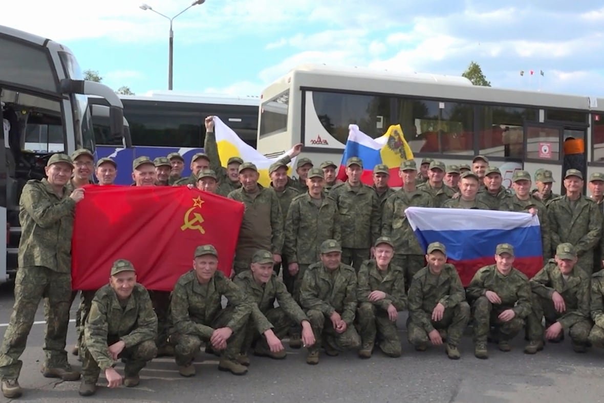 russia-ukraine-prisoners Dozens of military members in camouflage style uniform pose with flags. Some are crouching, some standing.