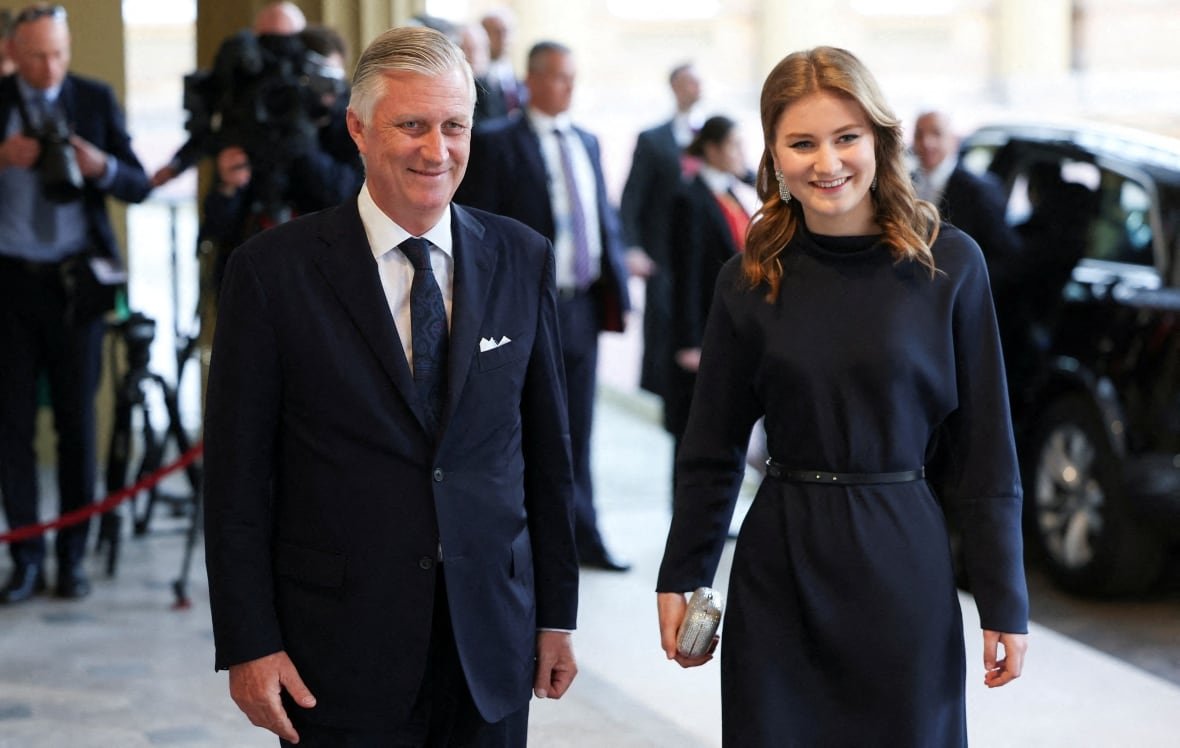 A white haired man in a suit and tie and a young woman are shown smiling and standing.