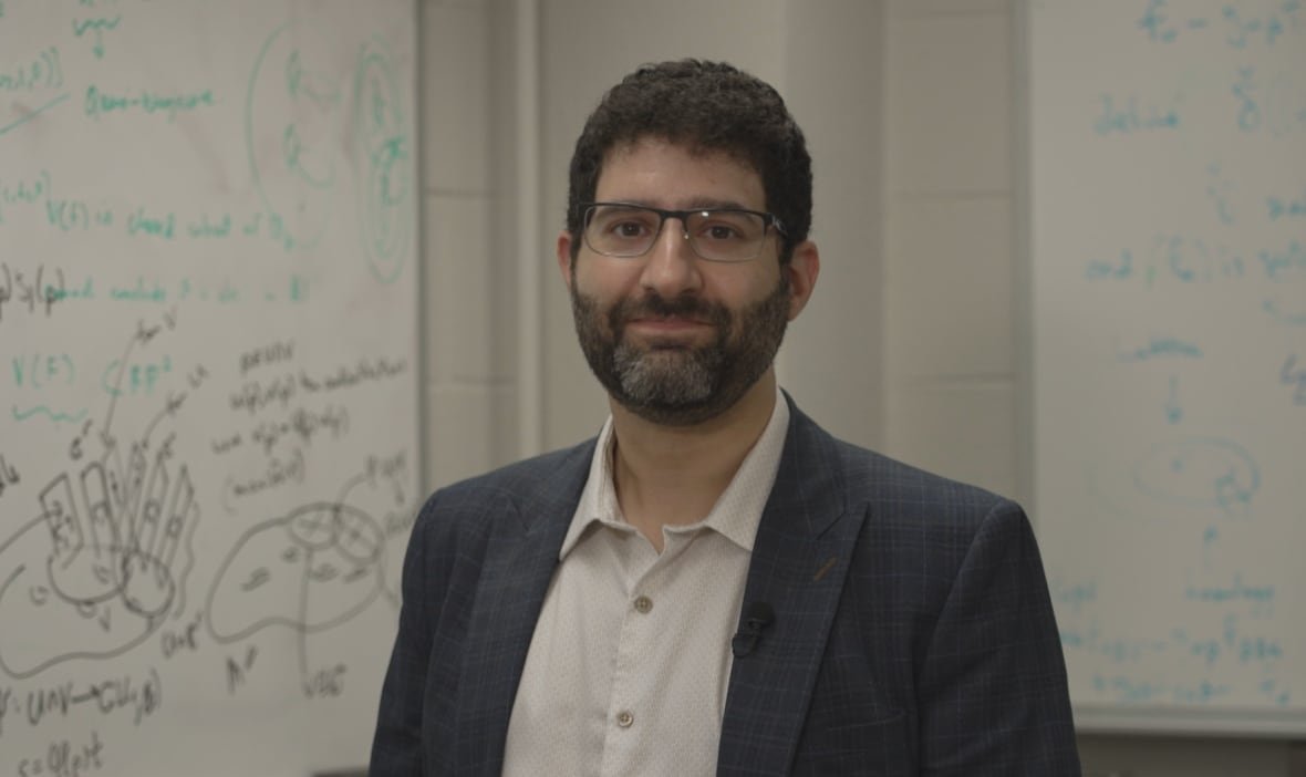 steven-rayan A man wearing glasses and a suit poses in front of a white board.