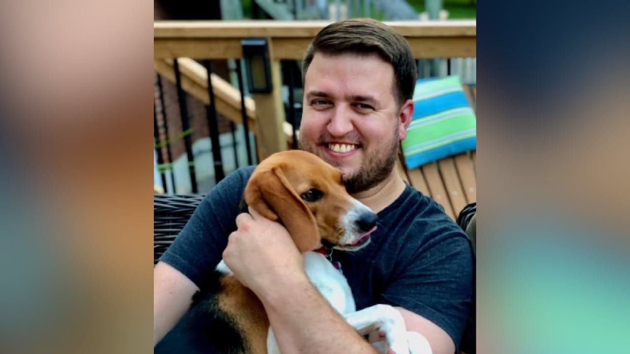 A man with dark hair and a dark beard smiles while looking at the camera. He's holding a beagle.