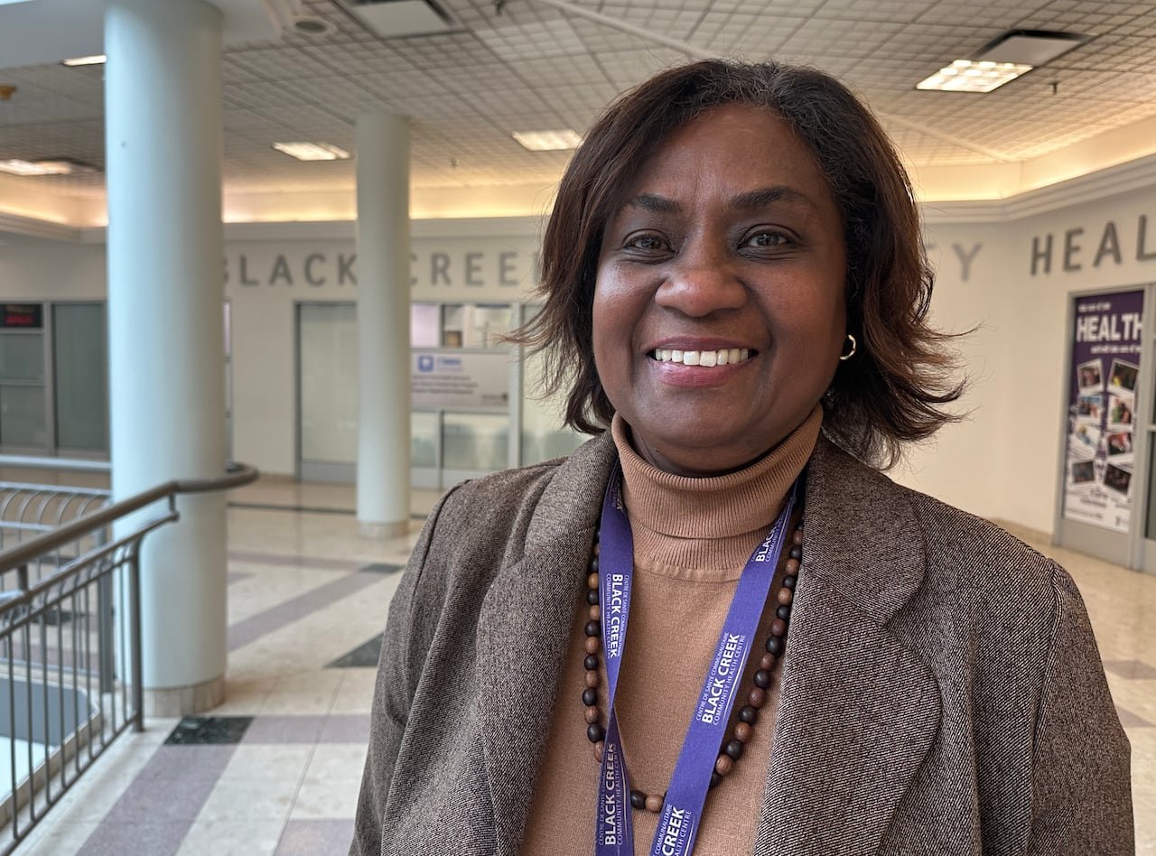 A woman smiles wearing a brown blazer, turtleneck, brown beaded necklace and purple lanyard.