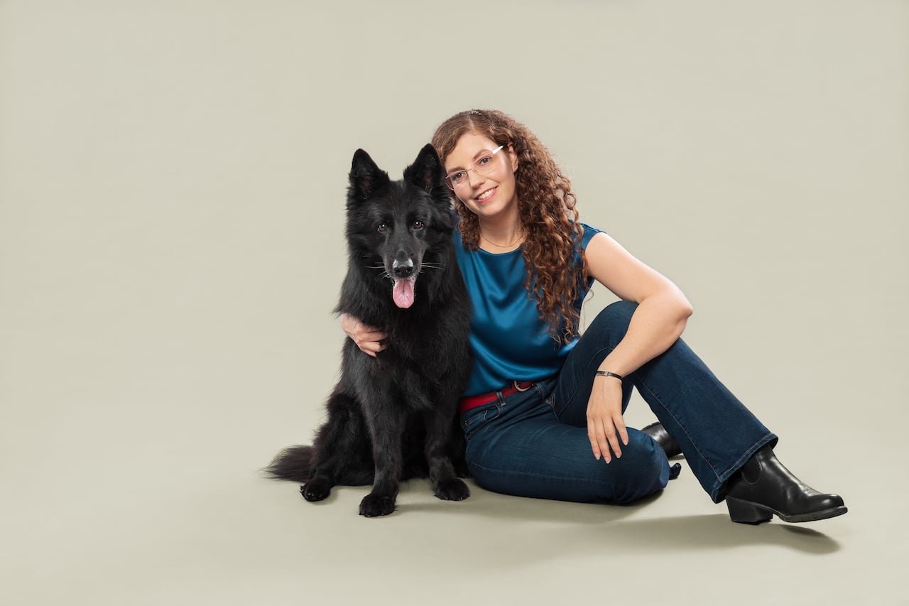 A woman sits smiling beside her large black dog