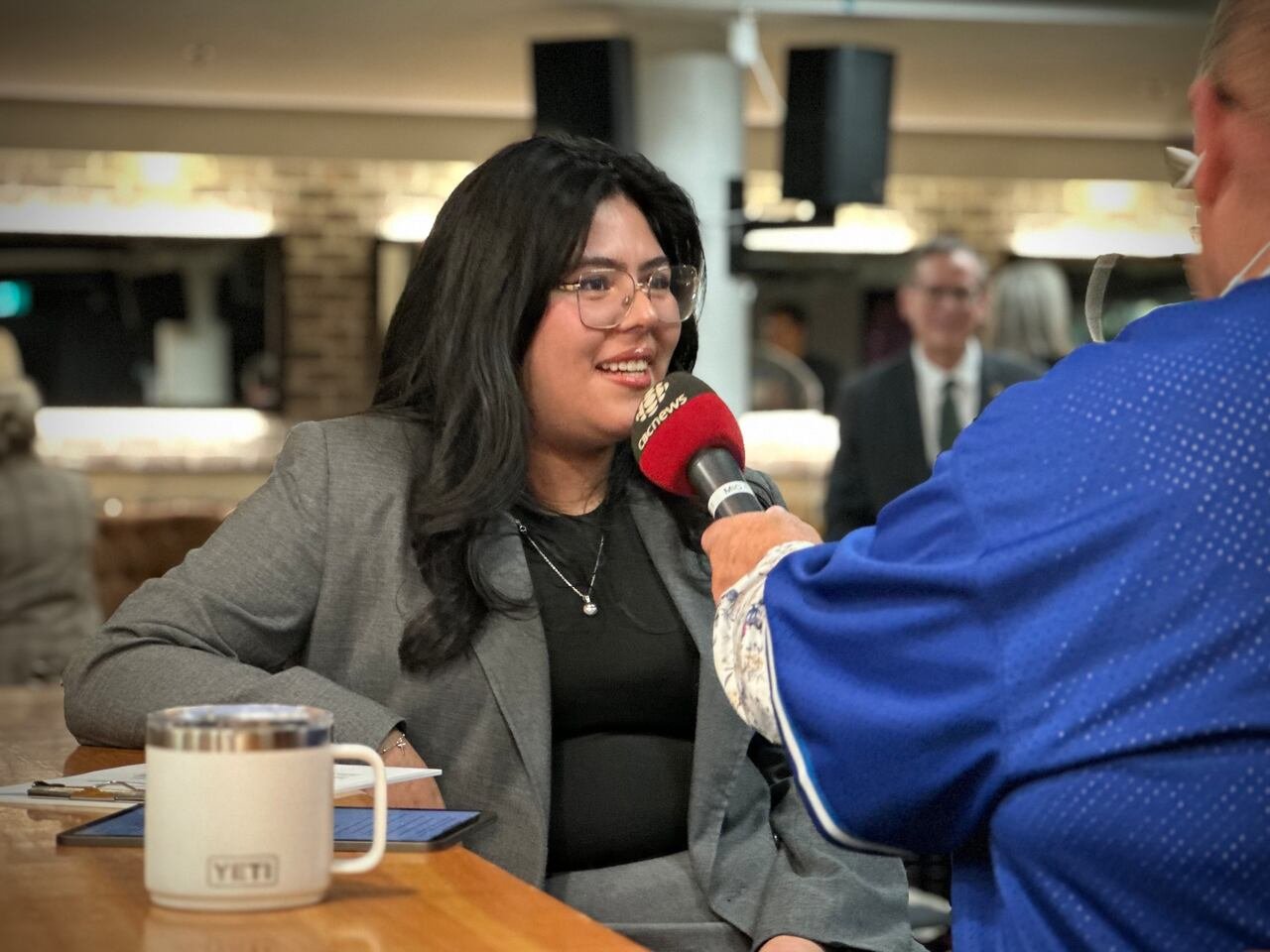 A woman with dark hair and glasses smiles while being interviewed by a reporter holding a CBC News microphone