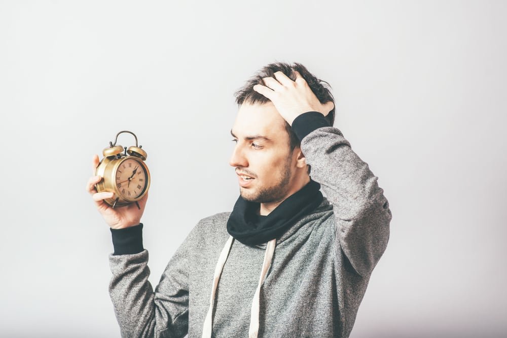 A man wearing a gray hoodie looks exasperated as he holds up an alarm clock.