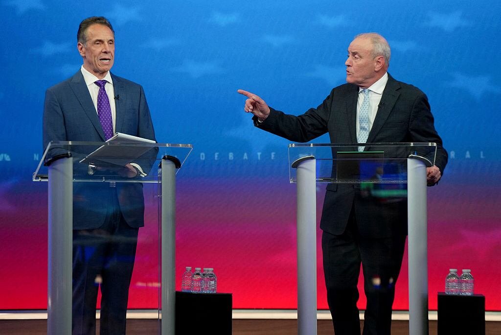 Two men in suits stand on a stage behind podiums. The man on the right gestures at the other man.