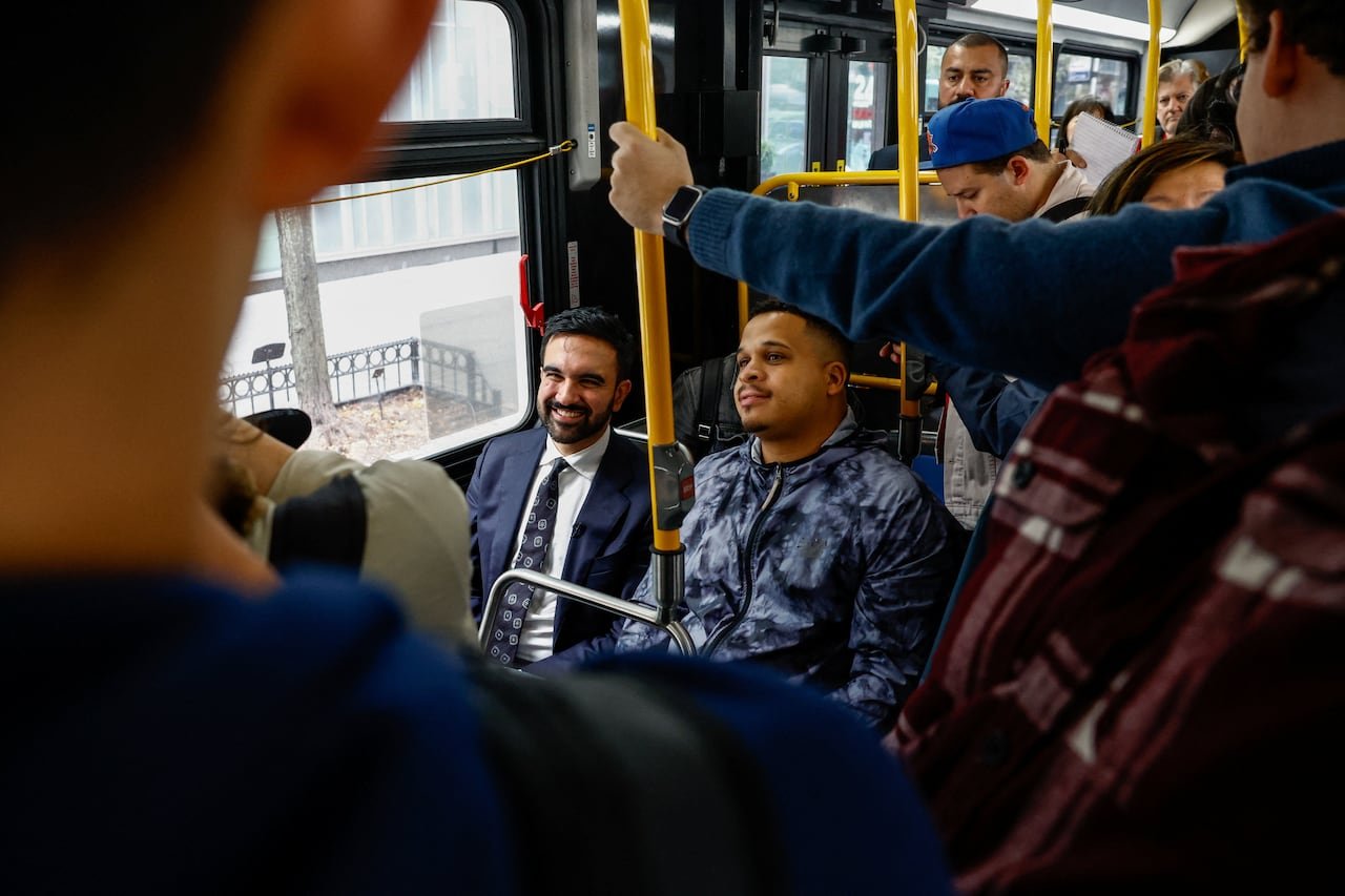 A man in a blue suit sits on a bus, smiling as riders look on.