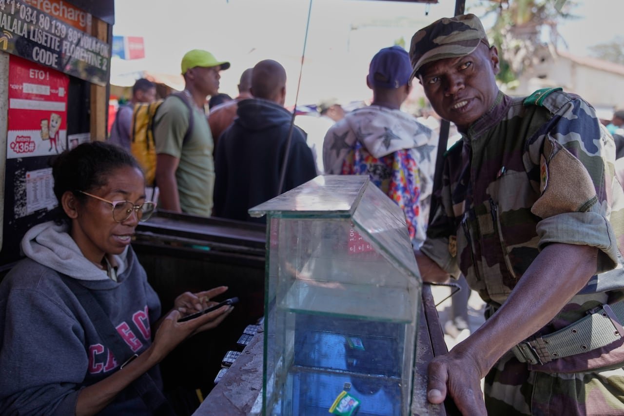 default-818 A man in military attire stands beside a seated woman wearing glasses in a market setting.