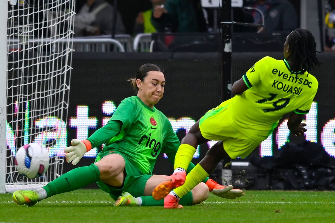 A female soccer player representing the Washington Spirit plays the ball past a goalkeeper representing the Portland Thorns.