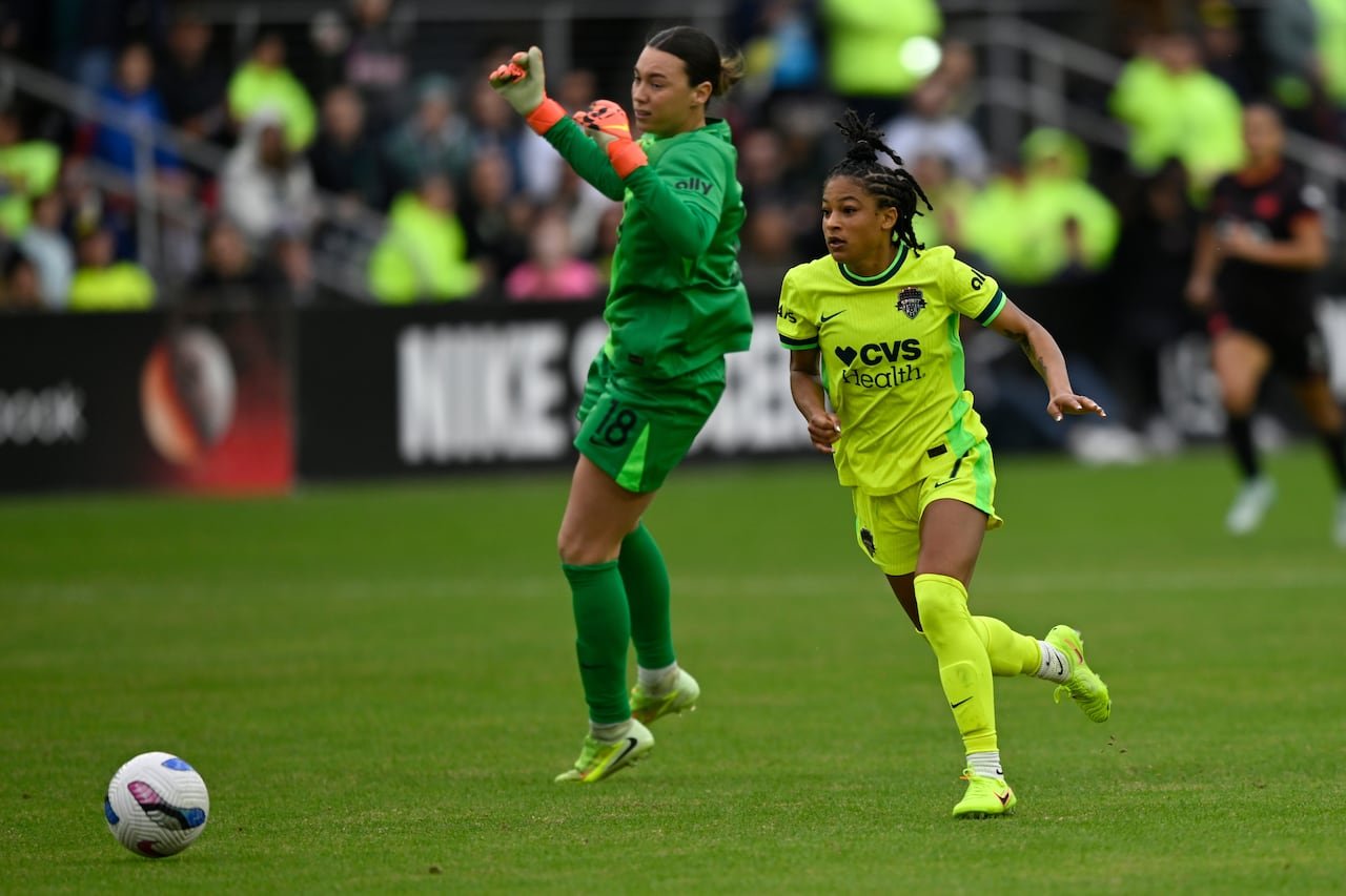 A female soccer player in a bright yellow and green uniform gets past an opposing goalkeeper.