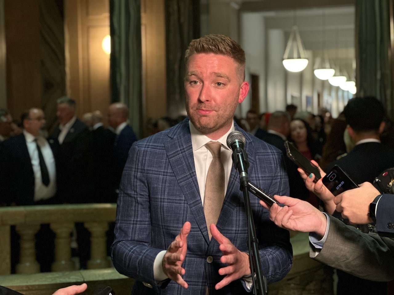 A man in a blue suit speaks into a microphone. The man is surrounded by hands holding recorders.