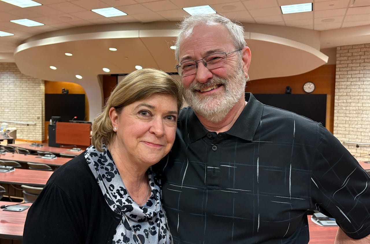 A woman and man in black stand close and smile for the camera inside a lecture hall