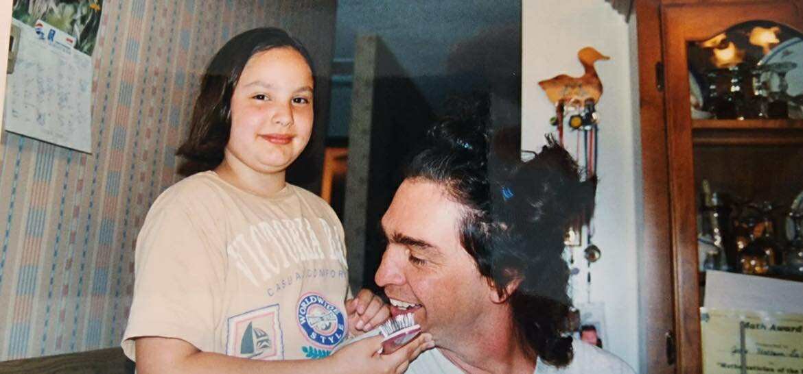 A child holding a hair brush stands beside a man with shoulder-length hair styled in ponytails.