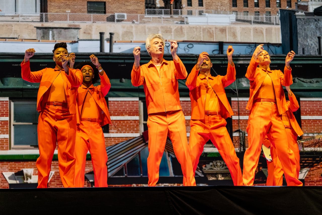 A male performer in an orange outfit with five backup singers dressed similarly performing on stage.