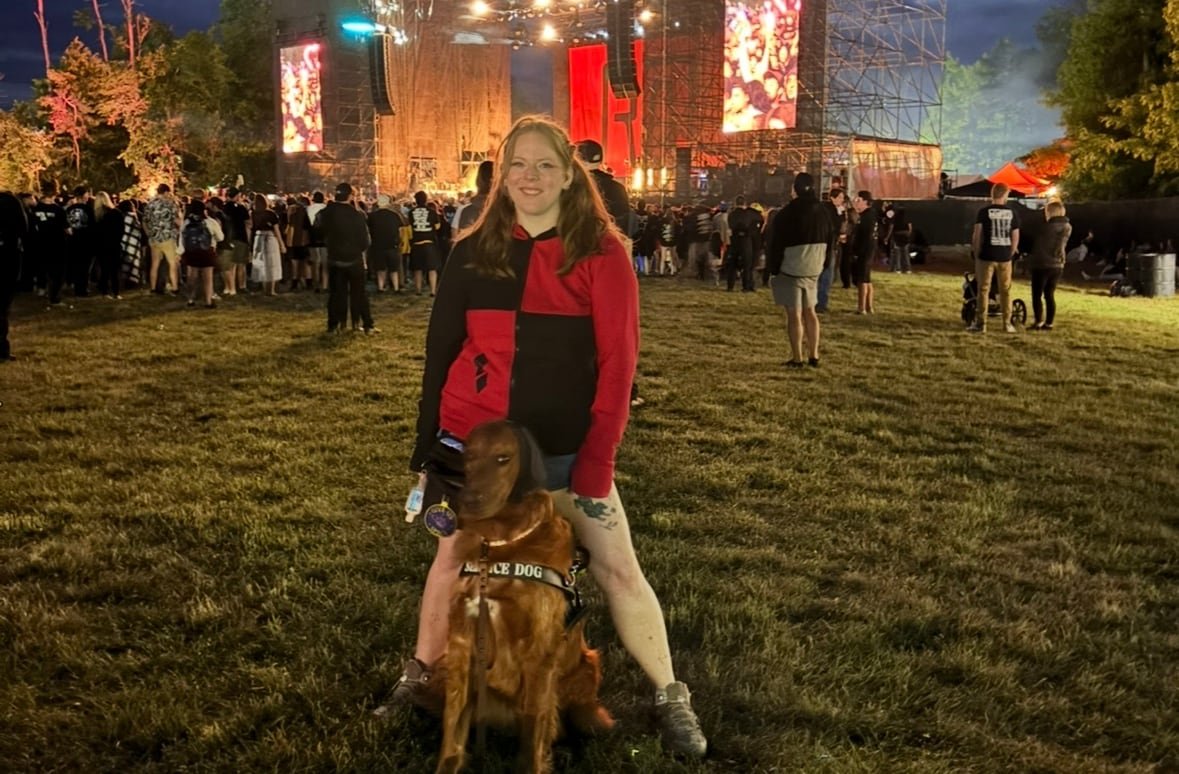 default-130 A woman stands in the crowd at an outdoor concert, her golden retriever standing between her legs.