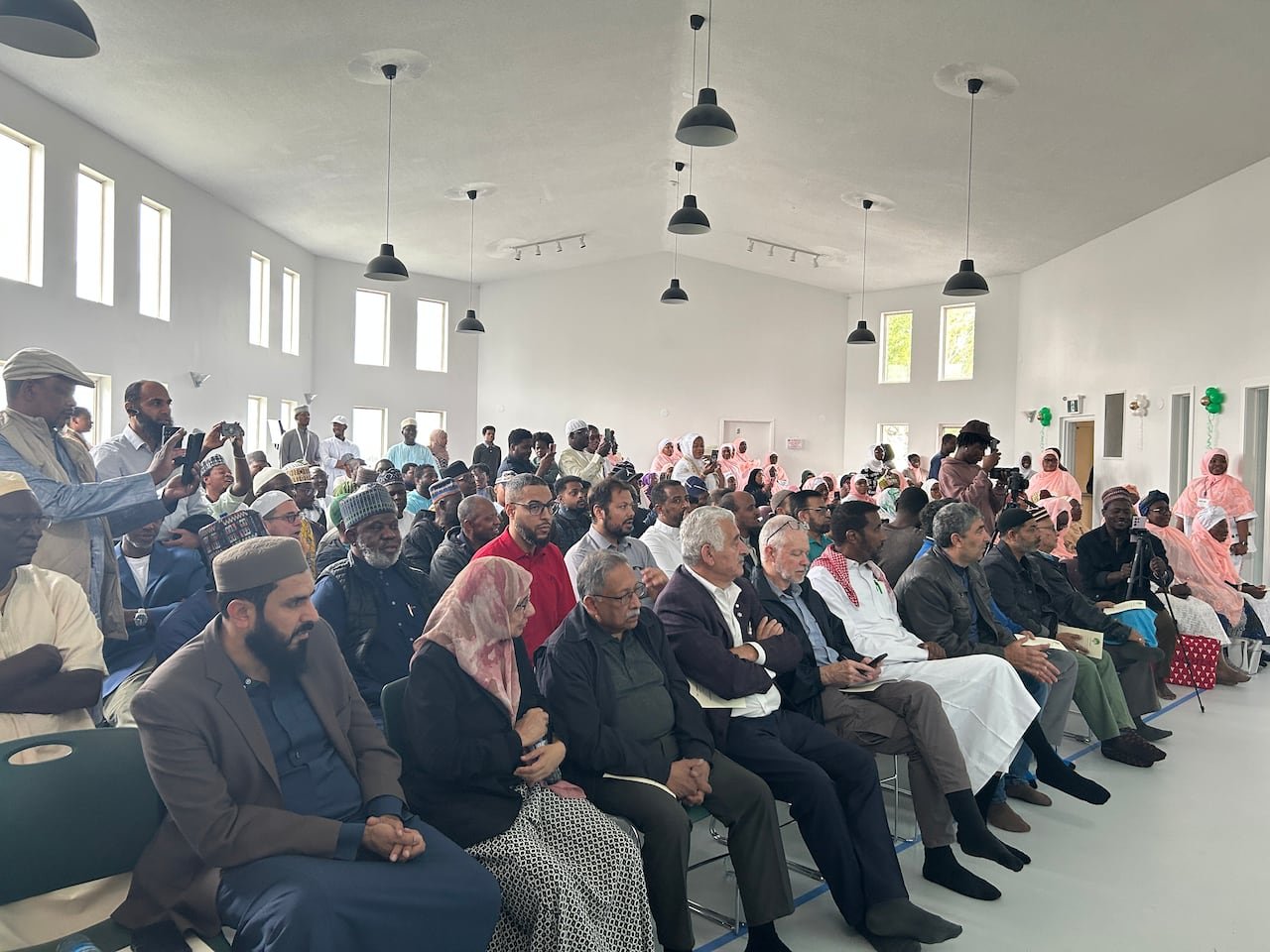 A crowd of dozens of people are seen in a large white room with windows