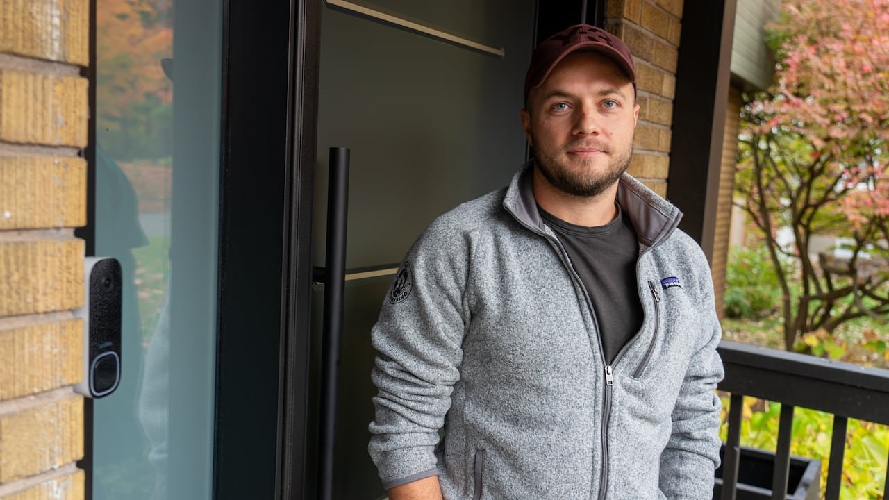 A man in a grey zip up sweater stands on his front porch.
