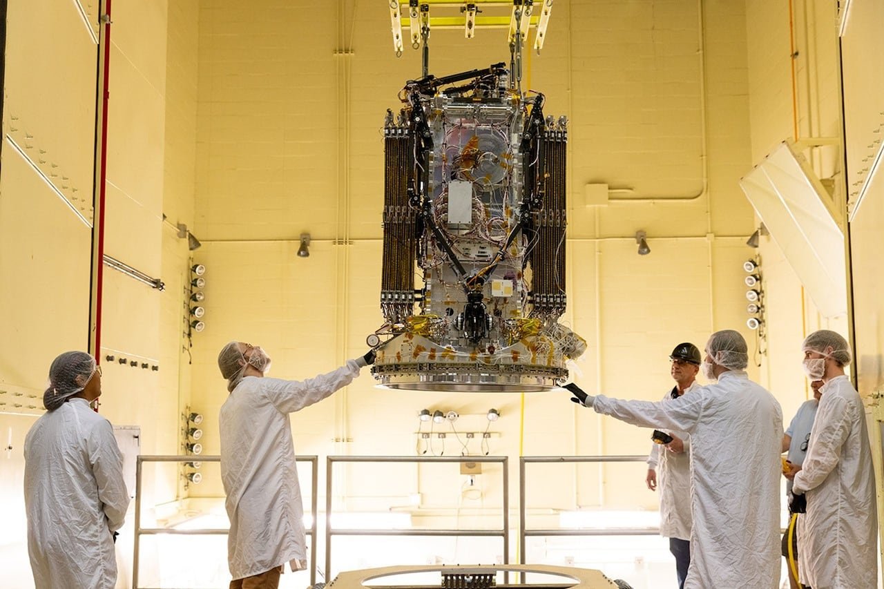 Men in white lab coats stand around a large cylindrical piece of technology hanging from a ceiling