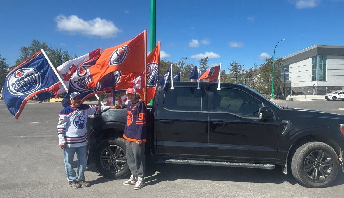 Two individuals in Edmonton Oilers jerseys stand beside a black truck adorned with multiple large Oilers flags.