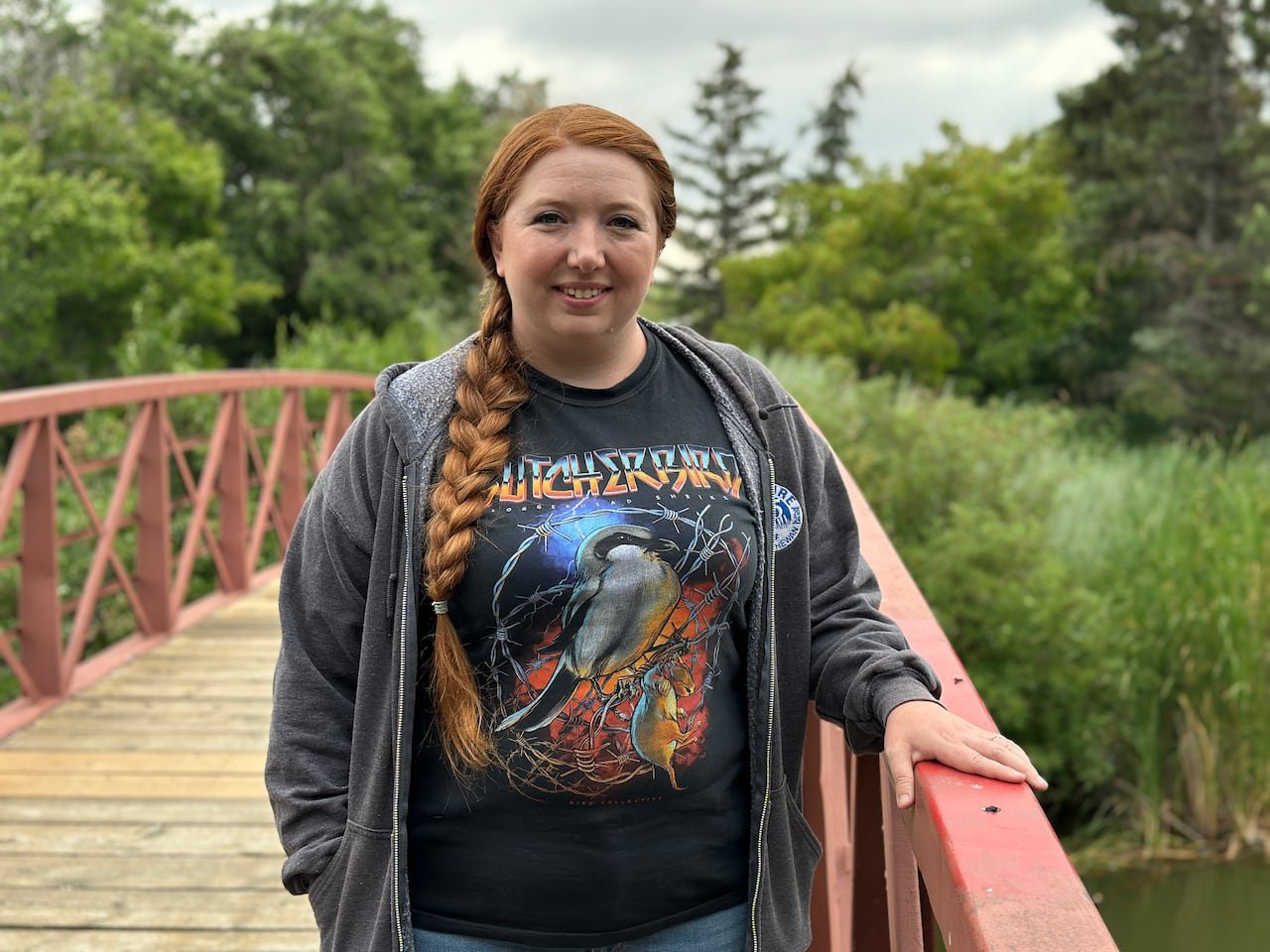 default-188 A woman with a long braid stands on a small bridge crossing a creek, smiling at the camera. She is wearing a shirt with a graphic depicting the "butcher bird," or loggerhead shrike.